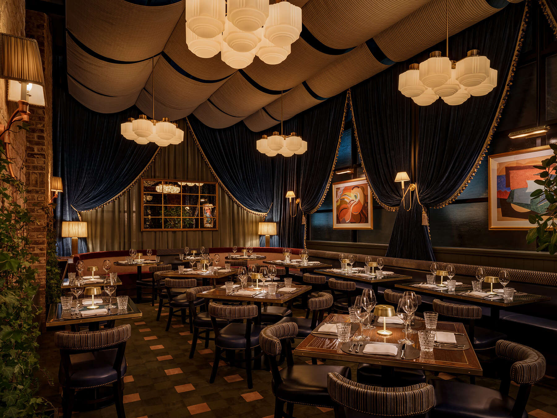 Stylish dining space with dramatic draped ceiling fabric, modern chandeliers, dark blue curtains, and neatly arranged tables set for dinner with glassware and ambient lighting.