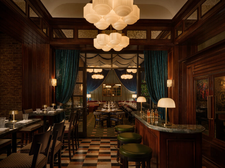 Refined restaurant bar area with rich wood paneling, green velvet stools, soft lighting, and a view into a dining room framed by tall curtains and elegant decor.
