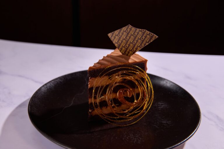 Slice of chocolate layer cake with caramel frosting and decorative chocolate garnish served on a black plate.