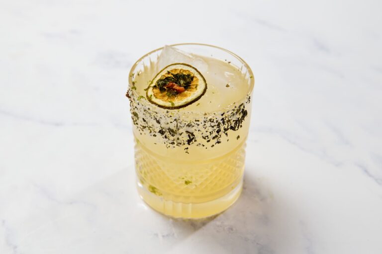 Yellow citrus cocktail with herb rim and dried citrus garnish served over ice in a rocks glass.