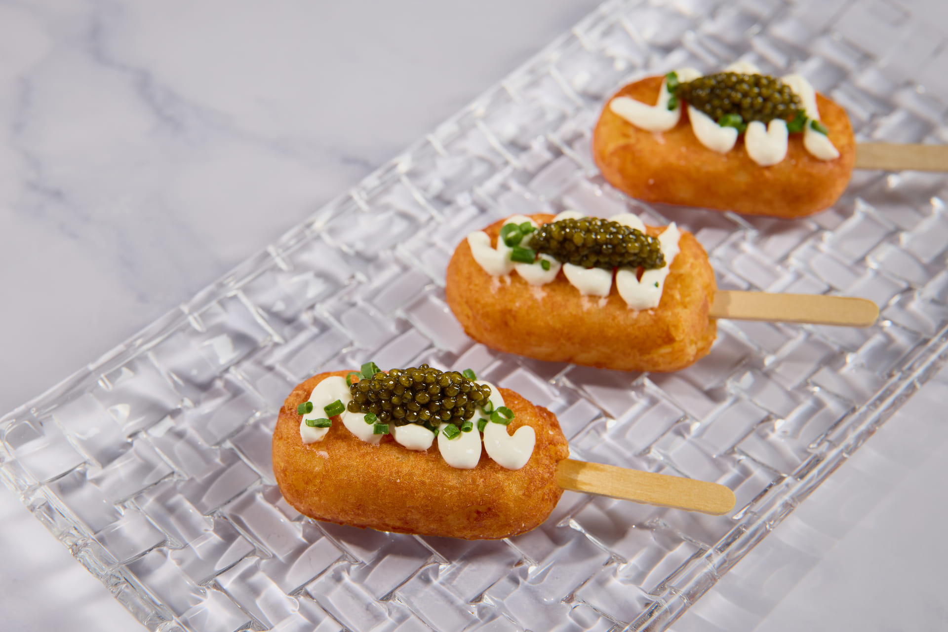 Mini corn dogs topped with crème fraîche, caviar, and chives served on a glass tray.