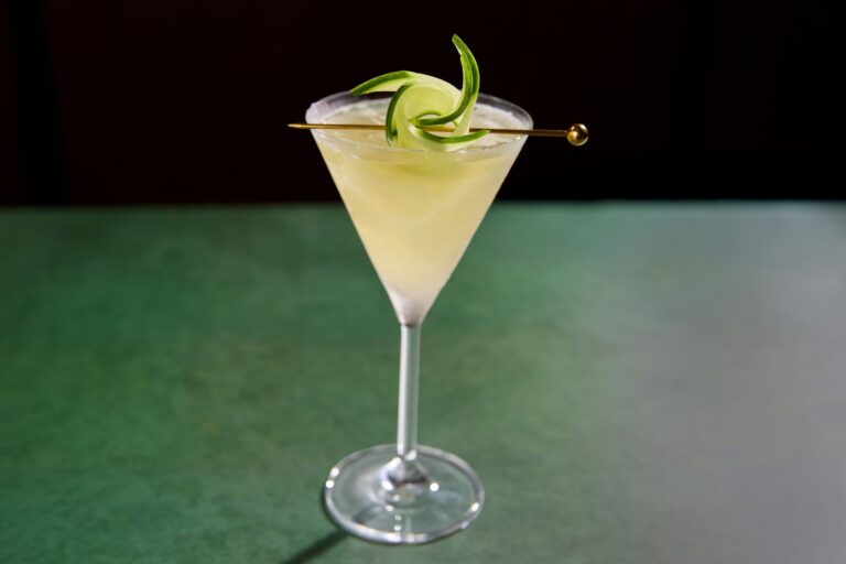 Pale green martini cocktail garnished with a curled cucumber ribbon in a martini glass.
