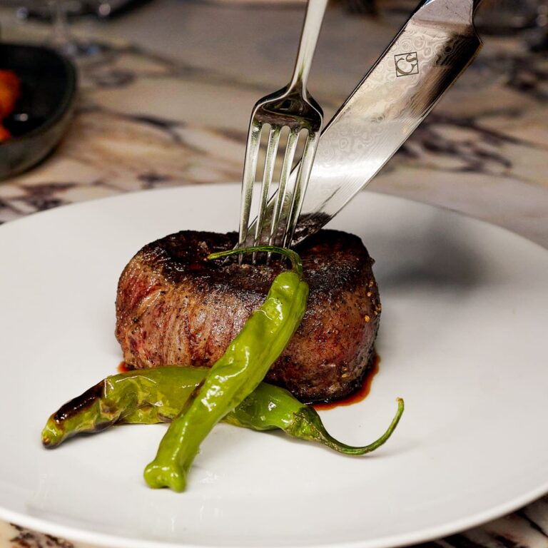 Grilled filet mignon topped with a fork and knife and served with blistered shishito peppers on a white plate.