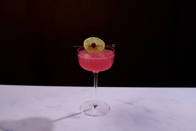 Pink cocktail served in a coupe glass garnished with a lime wheel and cherry.