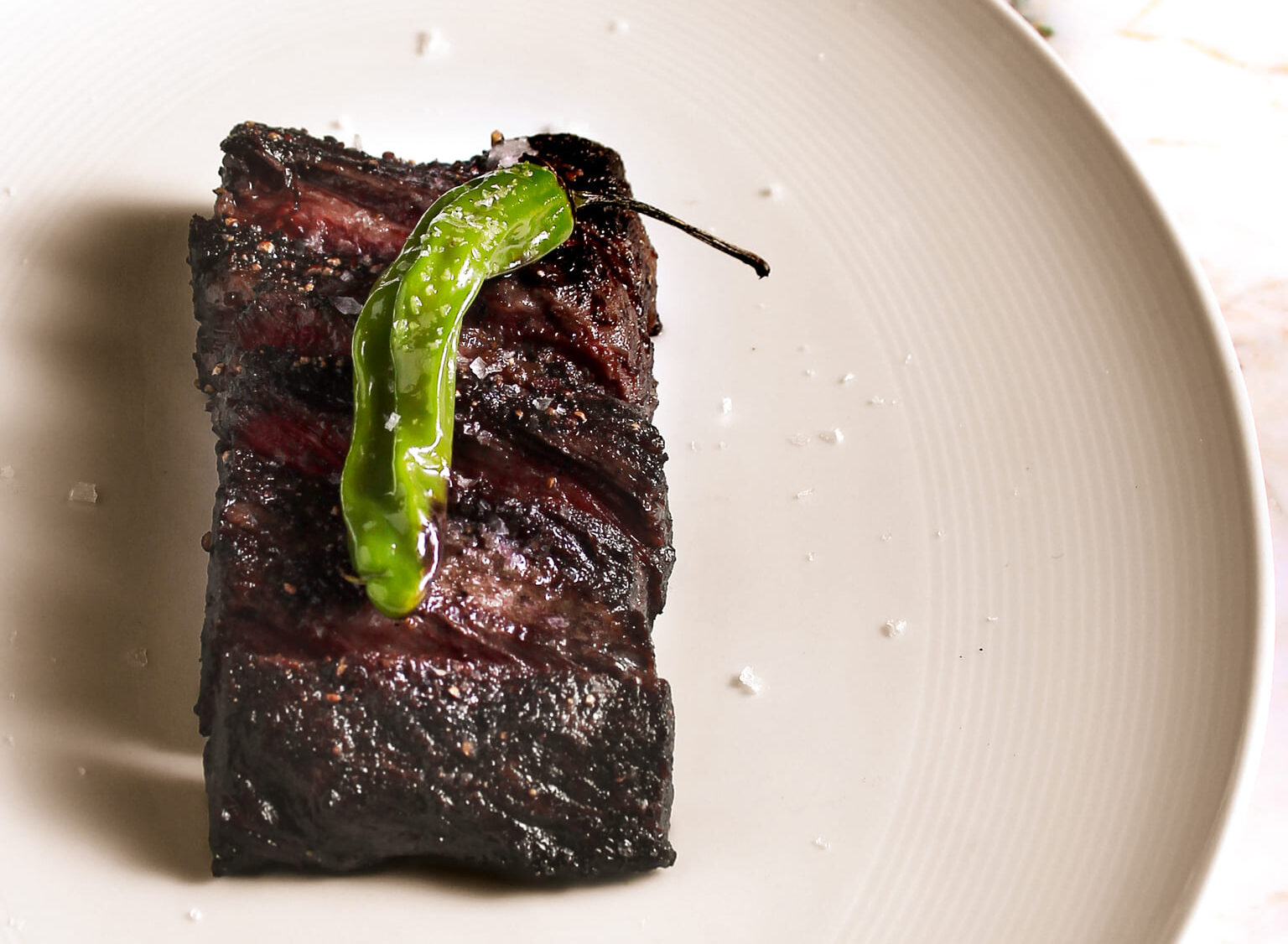Grilled rib cap steak topped with sea salt and a blistered shishito pepper on a white plate.