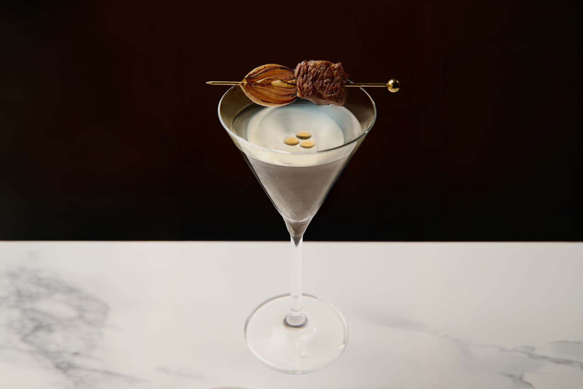 Clear martini cocktail garnished with grilled onion and a bite of beef on a skewer served in a martini glass.