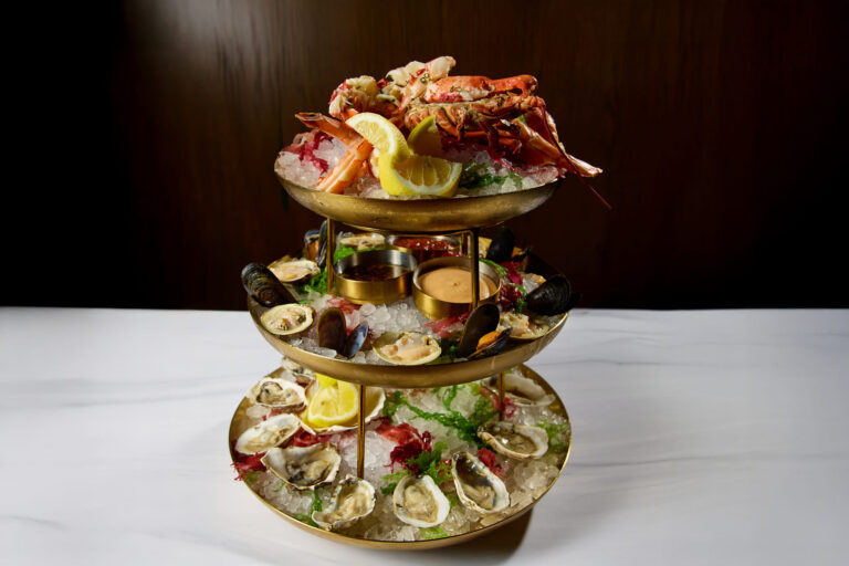 Three-tier seafood tower with oysters, mussels, lobster, shrimp, sauces, and lemon served on crushed ice.