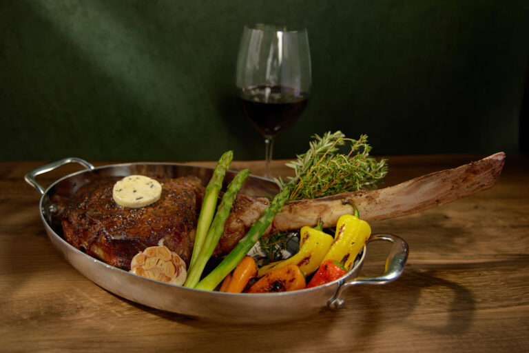 Tomahawk steak with herb butter served in a pan with asparagus, roasted peppers, garlic, and herbs.