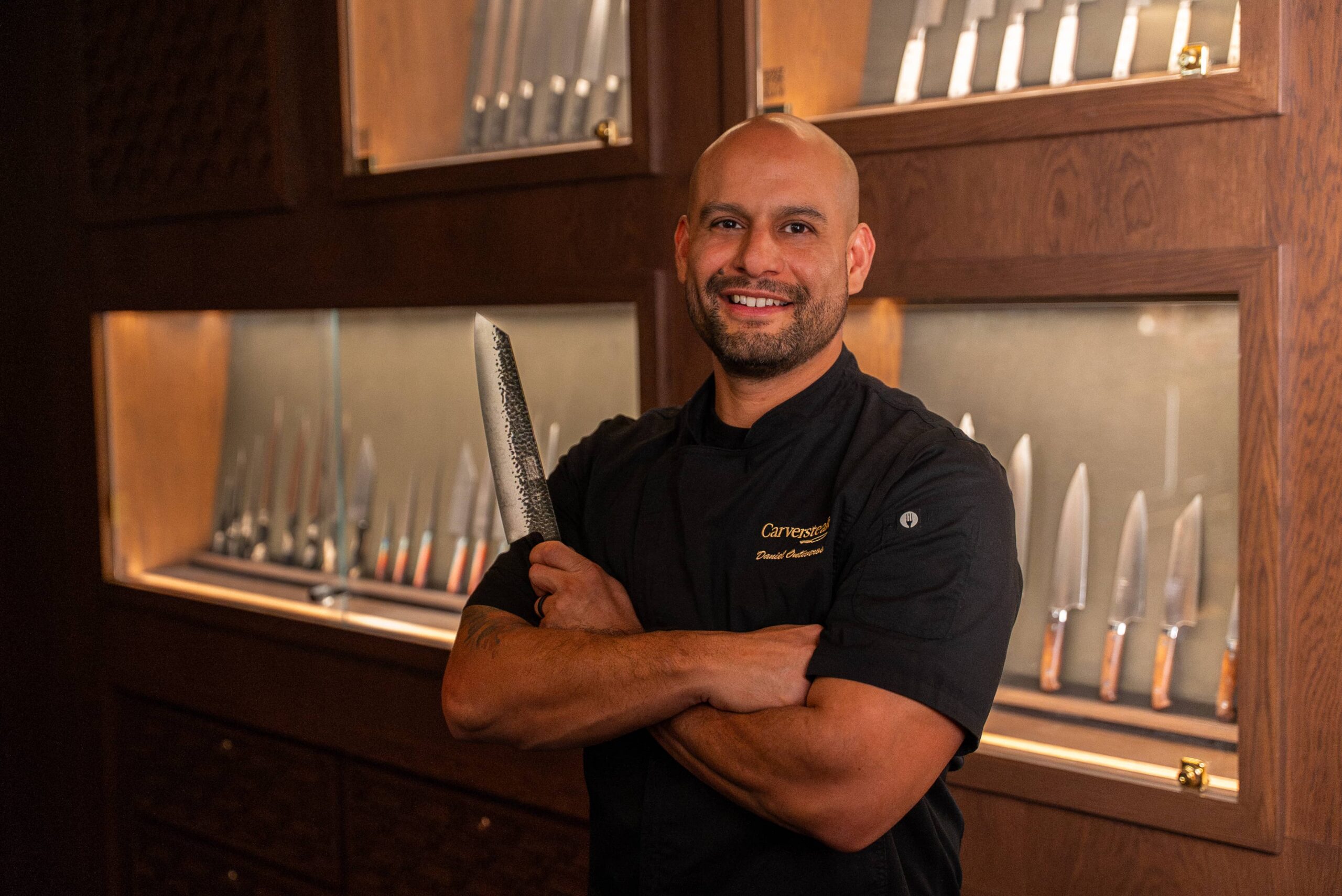 Portrait of a chef in a black uniform holding a chef’s knife, standing in front of a wall display of knives inside a restaurant kitchen.