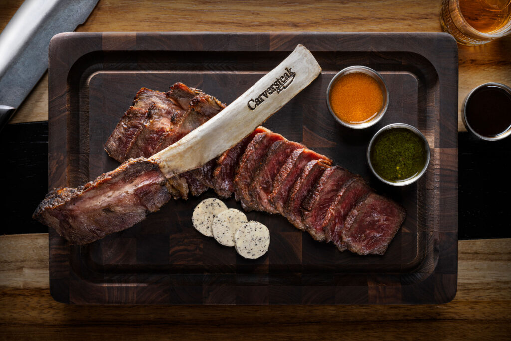 Tomahawk steak sliced and served on a wooden cutting board with compound butter and dipping sauces, featuring a branded bone and a chef’s knife on the side.