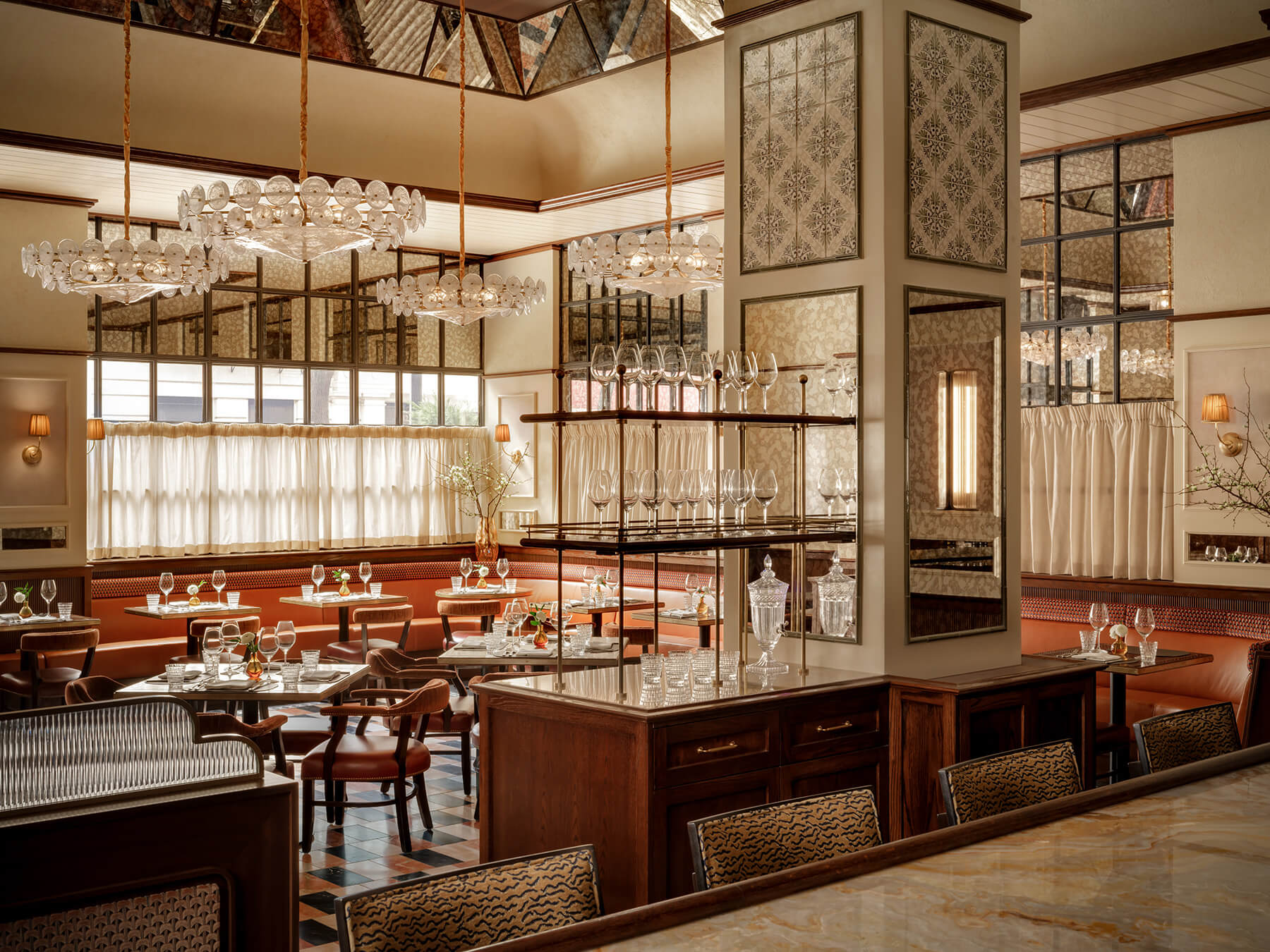 Bright and airy restaurant dining area with large windows, upholstered banquette seating, patterned tile floors, and tables set with glassware under decorative chandeliers.