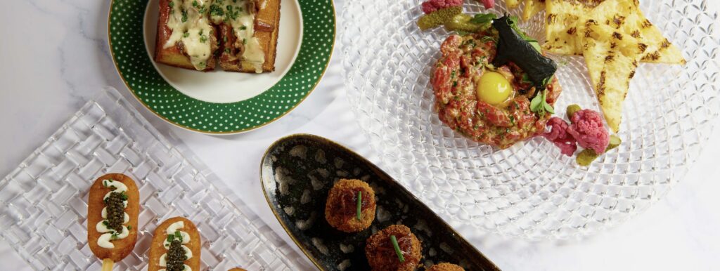 Overhead view of a gourmet spread featuring steak tartare with egg yolk and toasted bread, croquettes on a black plate, Caviar and Potato Popsicles, and a cheesy sandwich dish garnished with herbs on elegant tableware.