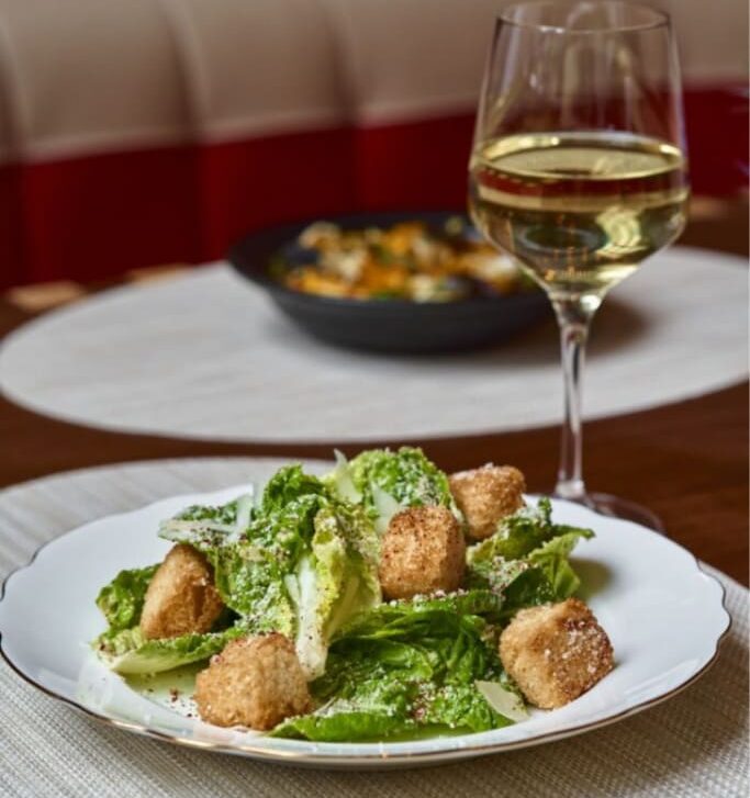 Caesar salad with croutons served on a plate alongside a glass of white wine at a restaurant