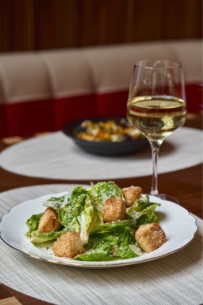 Caesar salad with croutons served on a plate alongside a glass of white wine at a restaurant