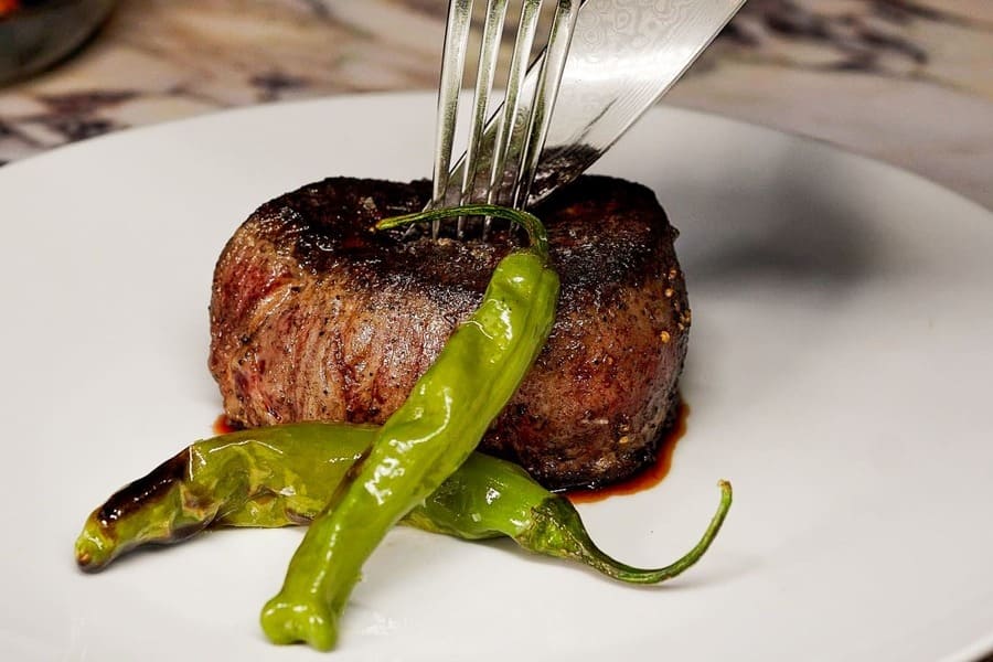 Close-up of a filet mignon steak on a plate with shishito peppers, being cut with a fork and knife.