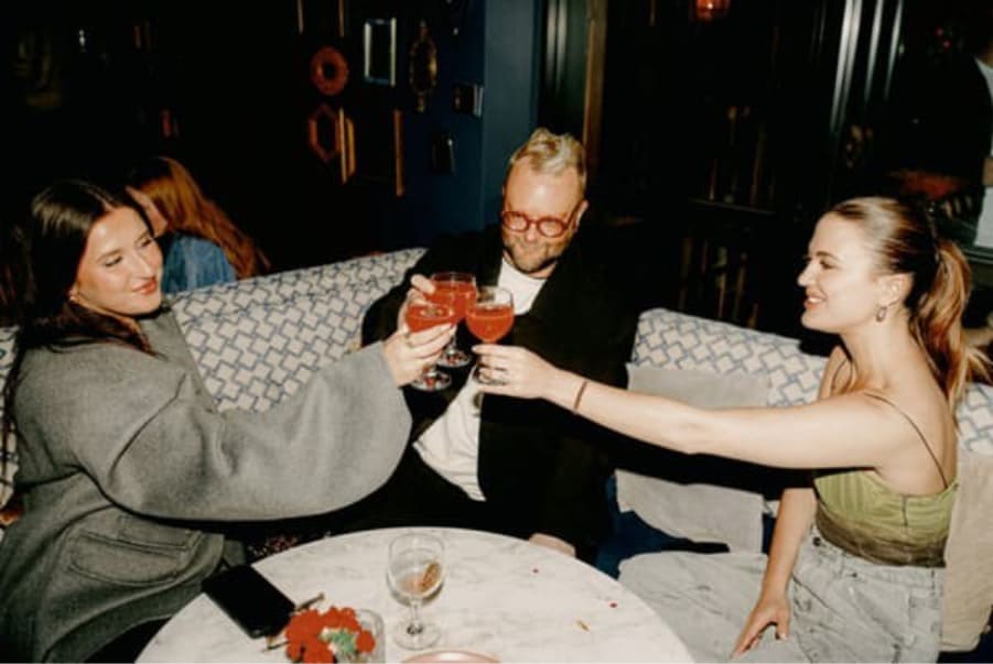 Three friends sitting in a lounge, smiling and toasting cocktails together at a table.