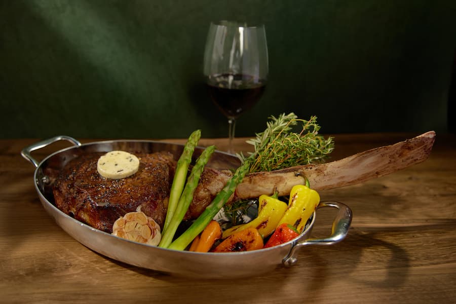 Tomahawk steak with herb butter served in a pan with asparagus, roasted peppers, garlic, and herbs.