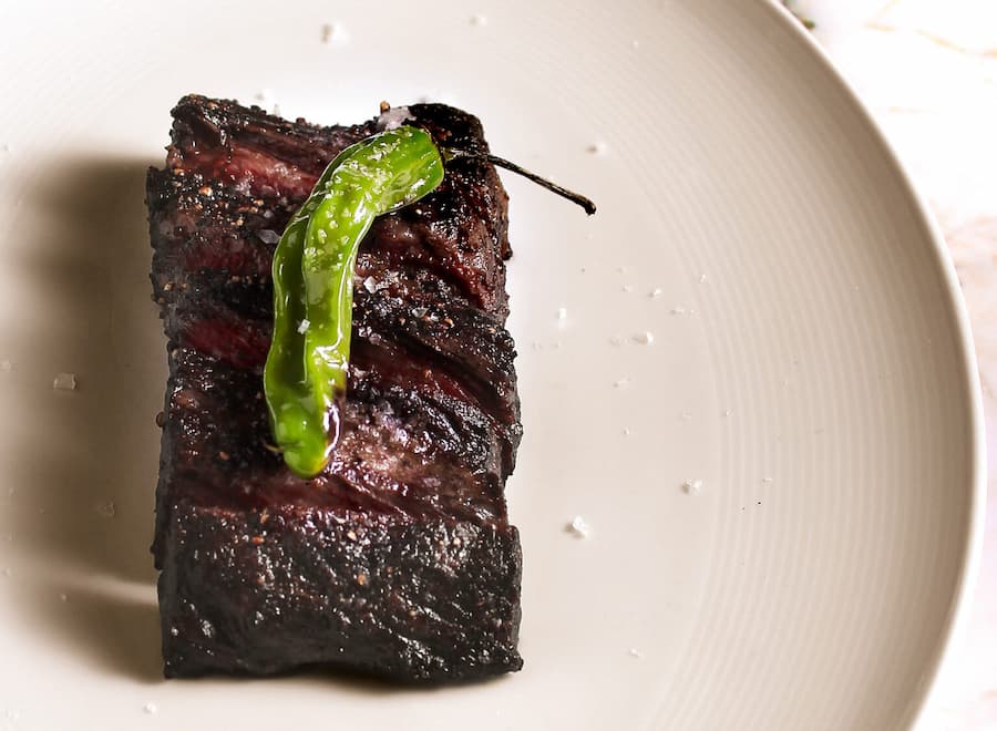 Grilled rib cap steak topped with a roasted shishito pepper and coarse salt on a white plate.