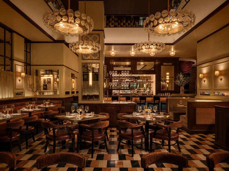 Elegant steakhouse dining room with warm lighting, chandeliers, wooden tables, and a bar in the background.
