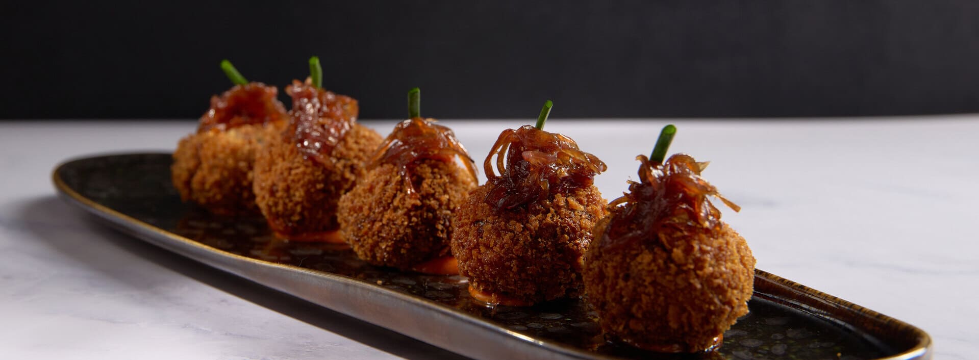 Five crispy braised short rib croquettes topped with caramelized onions and chives, served on a black rectangular platter with a savory sauce.