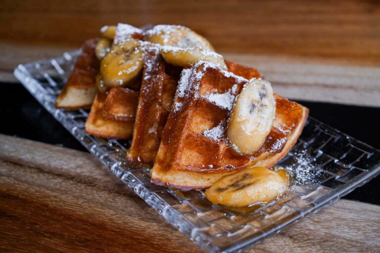 Golden waffles topped with sliced bananas, syrup, and powdered sugar on a glass serving plate.