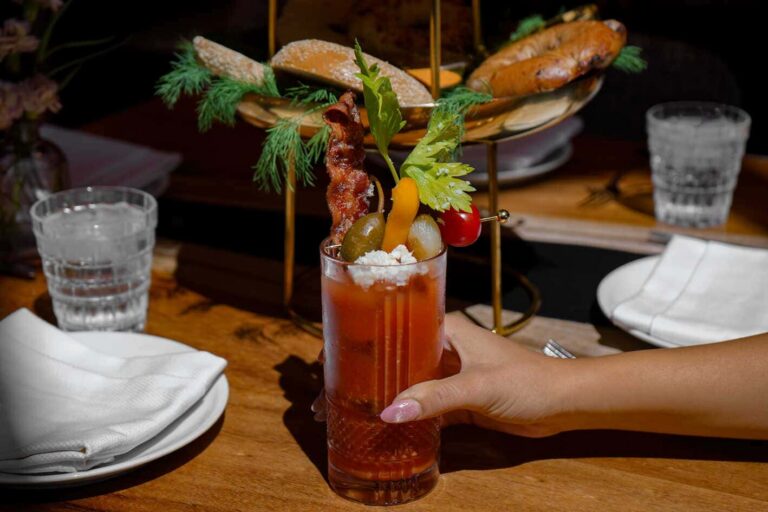 Bloody Mary garnished with bacon, celery, pickled vegetables, tomato, and cheese, served at a restaurant table.