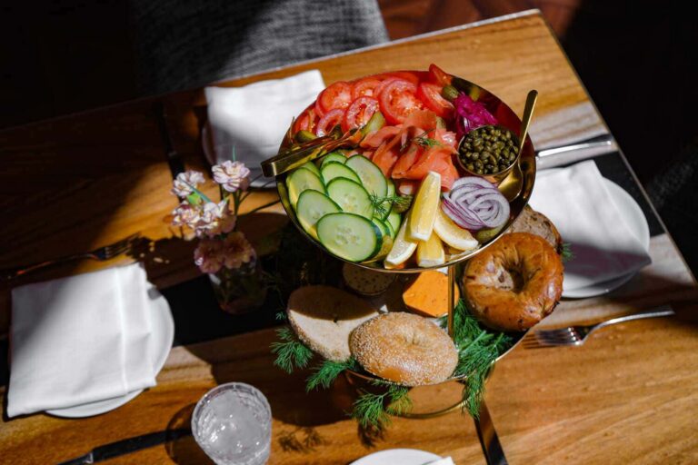 Brunch platter with smoked salmon, sliced tomatoes, cucumbers, capers, red onion, lemon wedges, and assorted bagels.