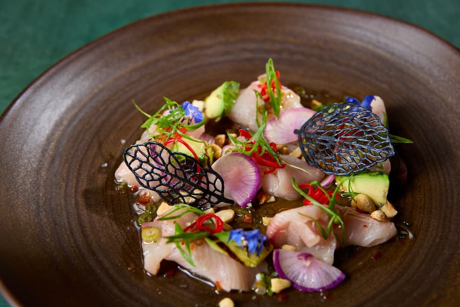 Hamachi crudo with avocado, radish slices, chili, herbs, and edible flowers served in a brown bowl.