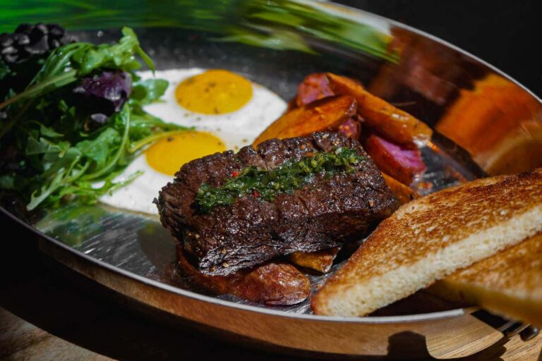 Grilled steak topped with herbs and served with two fried eggs, roasted potatoes, greens, and toast.
